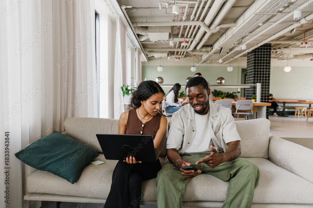 Obraz premium Male worker smiling and pointing at phone showing to female worker holding laptop while they sit on couch