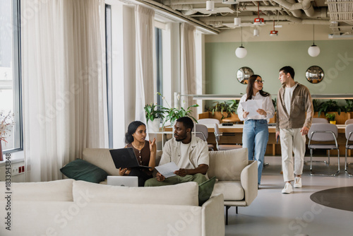 A female worker is holding a laptop and talking to a male worker who is listening to her and holding a document while they are sitting on a couch and two workers are walking towards them