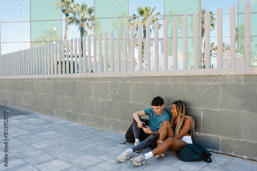 A male athlete is talking to a female athlete sitting next to him on the ground and laughing as they look at the phone he is holding and lean on the fence