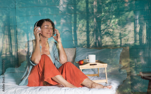 Mindfulness and feel-good moments.  lifestyle portrait smiling middle aged woman  in headphones rests while sitting on a bed near a window in a country house in the forest. Audio healing. 