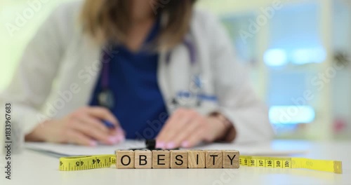 Wooden blocks spell word Obesity near measuring tape and pills while woman doctor writes notes. Woman specialist records data on weight problem