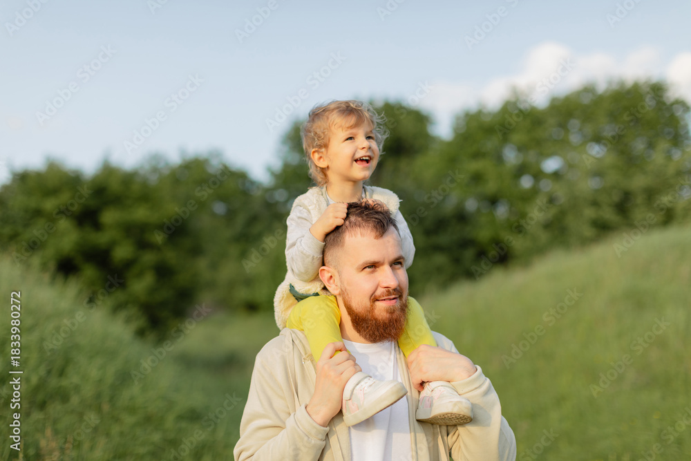 Fototapeta premium Father with a red beard carrying his toddler on his shoulders while walking outdoors on a sunny day. Warm natural light, joyful family moment and peaceful countryside atmosphere.
