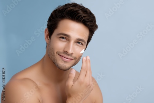 Handsome young man with smooth healthy skin is applying cosmetic cream to his face, showcasing a skincare routine in a bright and inviting atmosphere with soft blue background