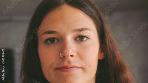 Close up shot of a young woman with brown hair wearing a silver nose ring with a subtle smile and