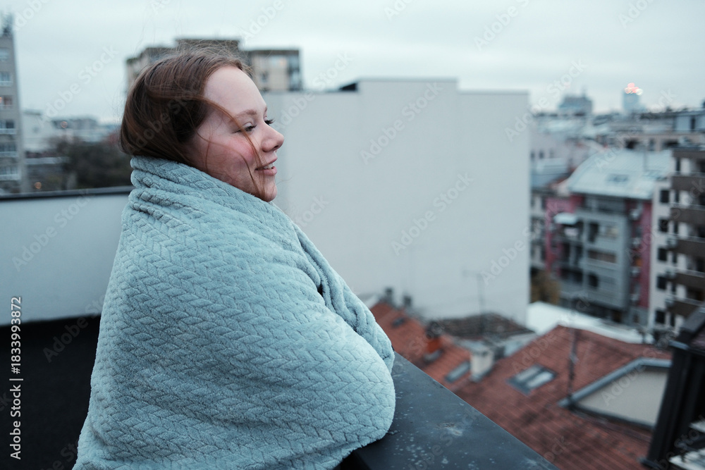 Obraz premium A woman wrapped in a soft blanket stands on a balcony and looks at the urban skyline. The concept expresses reflection and peaceful solitude.