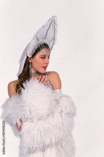 Elegant woman in an ornate white kokoshnik and luxurious faux fur coat posing gracefully in a clean studio setting. Sparkling accessories and soft lighting enhance her refined festive look