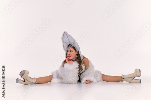 Stylish woman in a white kokoshnik and fluffy faux fur coat performing a full split in a clean studio setting, showcasing graceful flexibility and a striking festive performance look