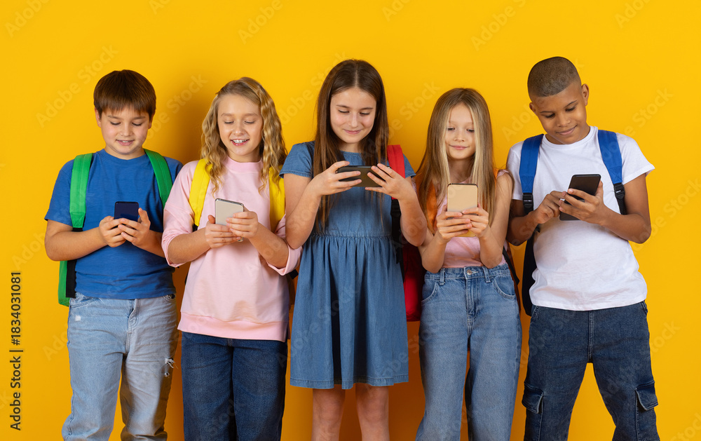 Obraz premium Preteen pupils standing together while looking at smartphones in bright studio background. A modern group of children engaged with technology in a school lifestyle scene