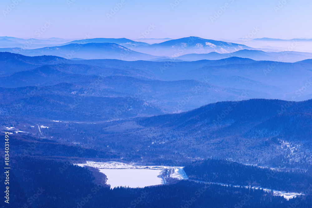 Fototapeta premium Panoramic view of mountain ridges near Sheregesh, Russia, covered in soft blue haze. Peaks fades into distance, creating depth effect under winter sky. Scenic landscape with frozen lake, reservoir.