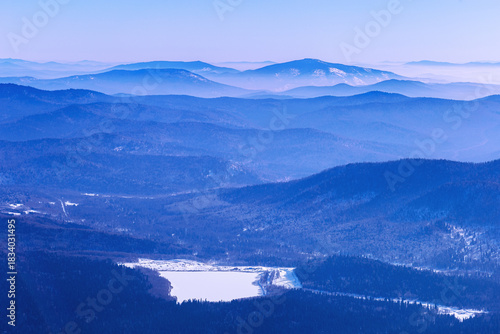 Panoramic view of mountain ridges near Sheregesh, Russia, covered in soft blue haze. Peaks fades into distance, creating depth effect under winter sky. Scenic landscape with frozen lake, reservoir.