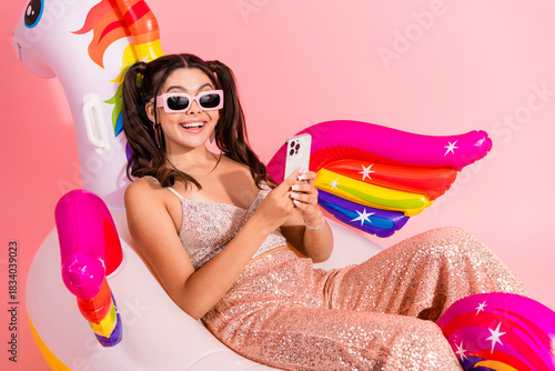 Young woman wearing a sparkly dress relaxes on a inflatable unicorn with rainbow wings in a pink studio smiles at her phone for a glamorous chic fashion lifestyle shot