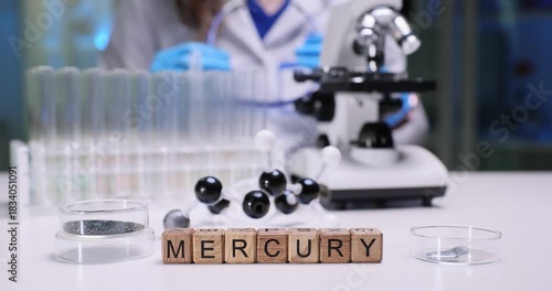 Word Mercury of wooden blocks against metal samples and molecular structure model on desk. Woman scientist checks goggles before tests start in lab