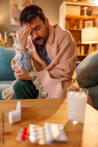 Sick man sitting on sofa wrapped in blanket having headache
