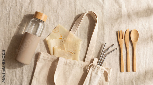 Sustainable living essentials arranged on a linen cloth include a water bottle, reusable wraps, utensils, and metal straws inside a cotton bag during the daytime.