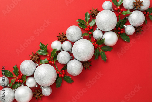 Christmas flat lay composition with white glitter baubles, holly berries, and leaves arranged diagonally on a vibrant red background. Festive holiday decor.