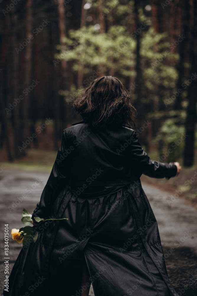 Naklejka premium Woman in a black coat walks down a forest path holding a yellow rose.