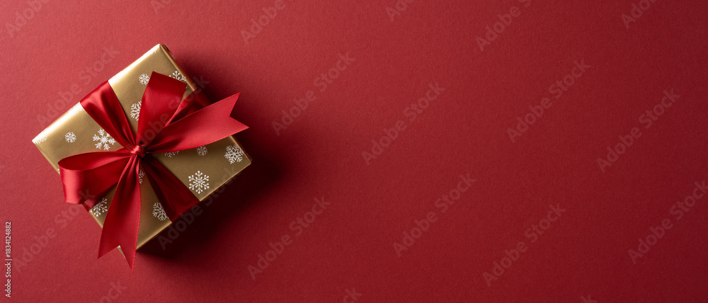 Fototapeta premium A gold wrapped gift box with a red bow rests on a deep red banner background, conveying festive mood, warmth, and celebration for Christmas and New Year