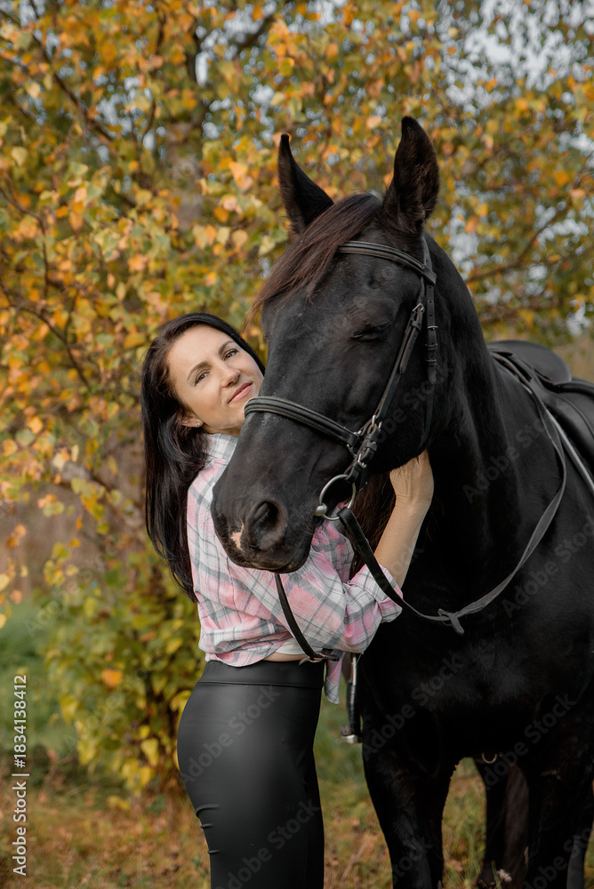 Obraz premium A young Caucasian woman with long dark hair hugs a large black horse in a colorful autumn setting with trees and fallen leaves.