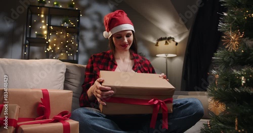 A woman wearing a Santa hat opens a Christmas gift box with a red ribbon, filled with anticipation and joy. A woman wearing a Santa hat is opening a Christmas gift on a couch, with a decorated tree