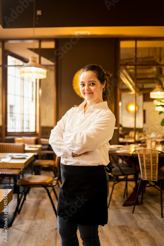 A young woman between 20 and 30 years old with Down syndrome works as a waitress in a modern cafe.The girl looks at the camera with her arms crossed happily.Concept of trisomy 21.
