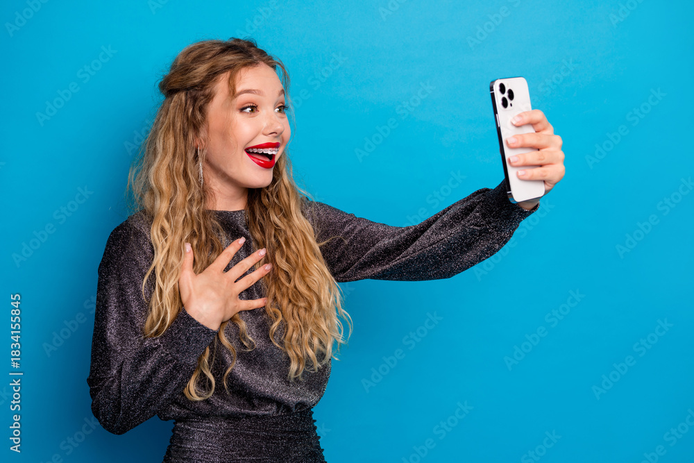 Fototapeta premium Joyful glamorous woman takes a selfie with a smartphone in a glittering dress against a bright blue background