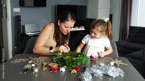 Mother and her little daughter decorating a Christmas wreath together at home. Strengthening family bonds through shared festive traditions and creative seasonal activities.