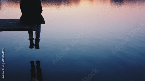 Silhouette Of Person Sitting On Dock Over Calm Water At Sunset. Reflective Moment Of Solitude