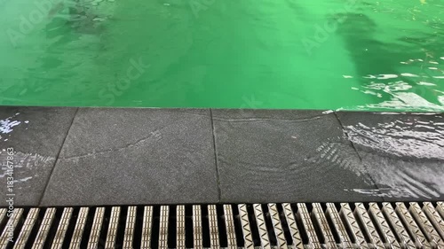 Vibrant green swimming pool water gently overflows a dark grey tiled edge into a drainage grate creating small ripples and a serene atmosphere