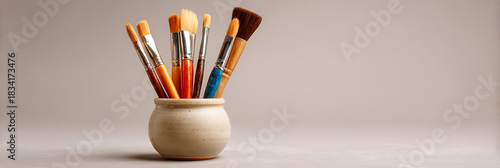 Collection of various artist paintbrushes in a ceramic jar isolated on a neutral beige background with copy space, representing creativity and art supplies.