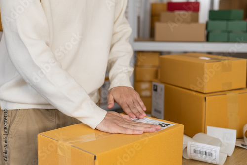 Warehouse shipping workflow showing young worker attaching label to parcel, preparing delivery orders, managing ecommerce packing process, supporting logistics operation in small business storehouse