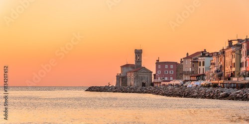 The coastal town of Piran in Slovenia  is illuminated by the rays of the setting sun over the sea. The coastal landscape is visible from the shore, with an orange sky.