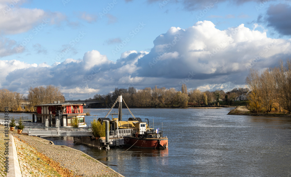 Fototapeta premium view of the Loire river along Quai du Chatelet in Orleans, France, during autumn. The paved promenade is lined under a dramatic cloudy sky with moored houseboats