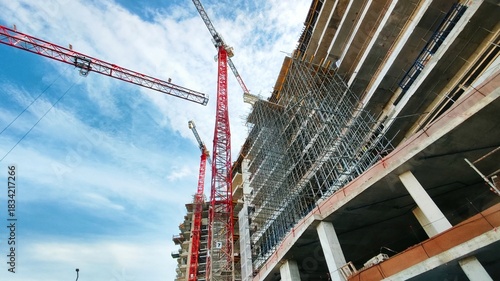 A ground-level worm's view of a construction site, looking up at scaffolding and cranes at the front of a building under construction