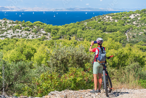 Young woman in sportswear with a mountain bike stands and takes photos, coastal landscape with sea and mountains, view from a hill with a gravel bike path on a sunny summer day.