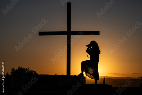 Silhouette of a cross and a young girl taking a photograph at sunset at the Grotto of Lourdes in Villa Giardino, Cordoba, Argentina.