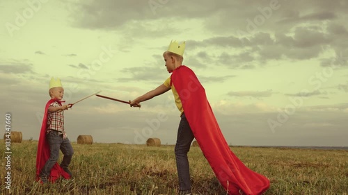children superheroes play with swords field kings knights. dream childhood. little kid and his brother fearless defenders. teamwork child boys sons. dream adventure red capes at sunset. summer games