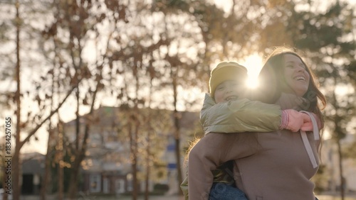 little child is having fun with her mother in city park, happy family life, kid girl on her back mom laughs, childish desires play, daughter love hug mom, laughing cheerful people on street glare sun.