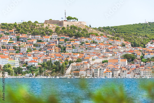 The coastal town of Sibenik in Croatia sits on the seafront, with hilltop houses and the Barone Fortress. A view from the other side on a sunny summer day.