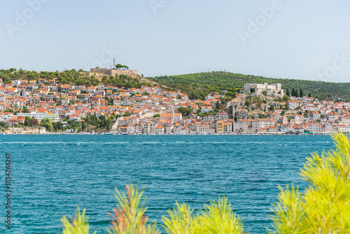 The coastal town of Šibenik in Croatia sits on the seafront, with hilltop houses, the Barone Fortress, and St. Michael's Castle. View from the other side on a sunny summer day.