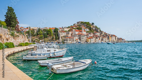 The coastal town of Sibenik in Croatia sits on the seafront, with hilltop houses and St. Michael's Castle. Boats moored along the shore, viewed from the walking path on a sunny summer day.