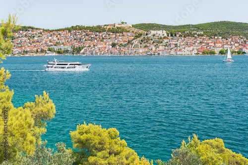 The coastal town of Šibenik in Croatia sits on the seafront, with hilltop houses, the Barone Fortress, and St. Michael's Castle. View from the other side on a sunny summer day.