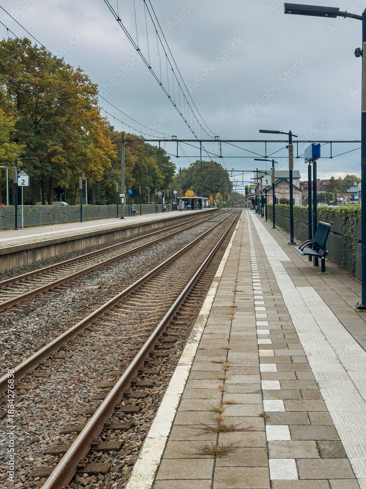 Fototapeta premium Dutch train station platform with a bench, tracks, overhead wires, and autumn trees receding into distance.