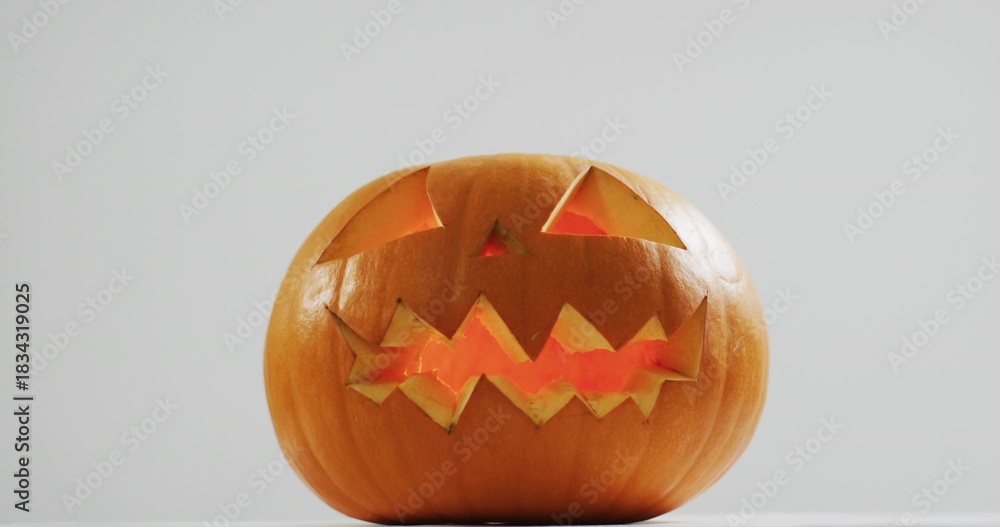 Fototapeta premium Glowing single carved pumpkin emitting warm candlelight on white platform against light-gray wall