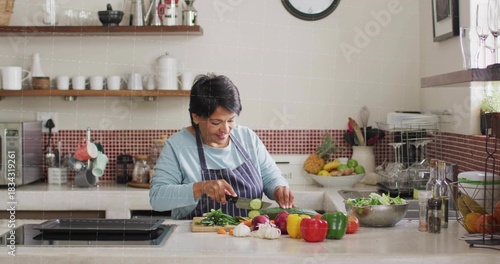 Chopping senior Indian cook in striped apron slicing cucumber at kitchen island, with cutting board
