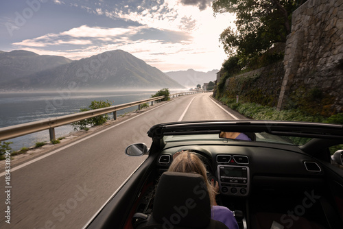 Young woman driving convertible along scenic ocean coast and mountain road at sunset, carefree summer vacation, travel freedom and adventure on coastal highway