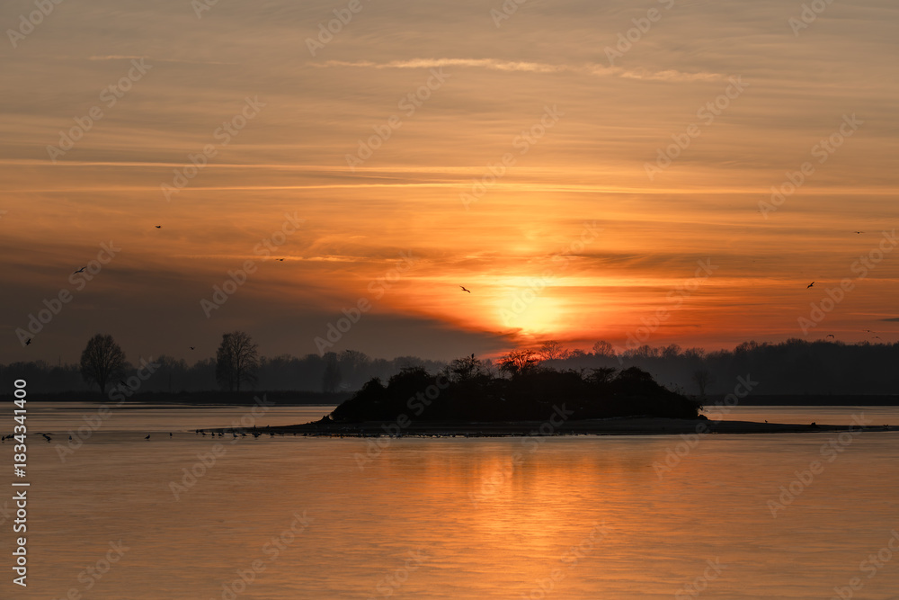 Naklejka premium Zachód słońca nad zamarzniętym stawem z sylwetkami ptaków stojących na lodzie. Ciepłe, wieczorne kolory kontrastują z zimną, teksturowaną powierzchnią zamarzniętej wody, tworząc spokojną i nastrojową