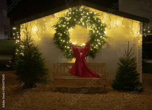 A large glowing Christmas wreath with a red bow decorates a small wooden hut, framed by fairy lights and festive evergreen trees.