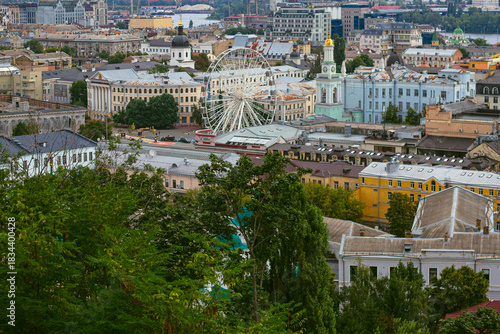 Kyiv Podil Seen from Andriyivskyi Descent with Kontraktova Square Wheel, Green Trees, Rooftops of Historic Buildings, and Distant Left-Bank High-Rises