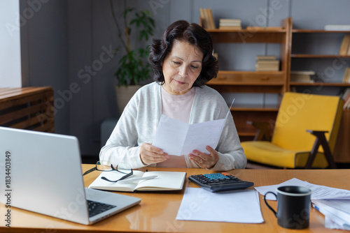 Middle aged senior woman sit with laptop and paper document. Pensive older mature lady reading paper bill pay online at home managing bank finances calculating taxes planning loan debt pension payment