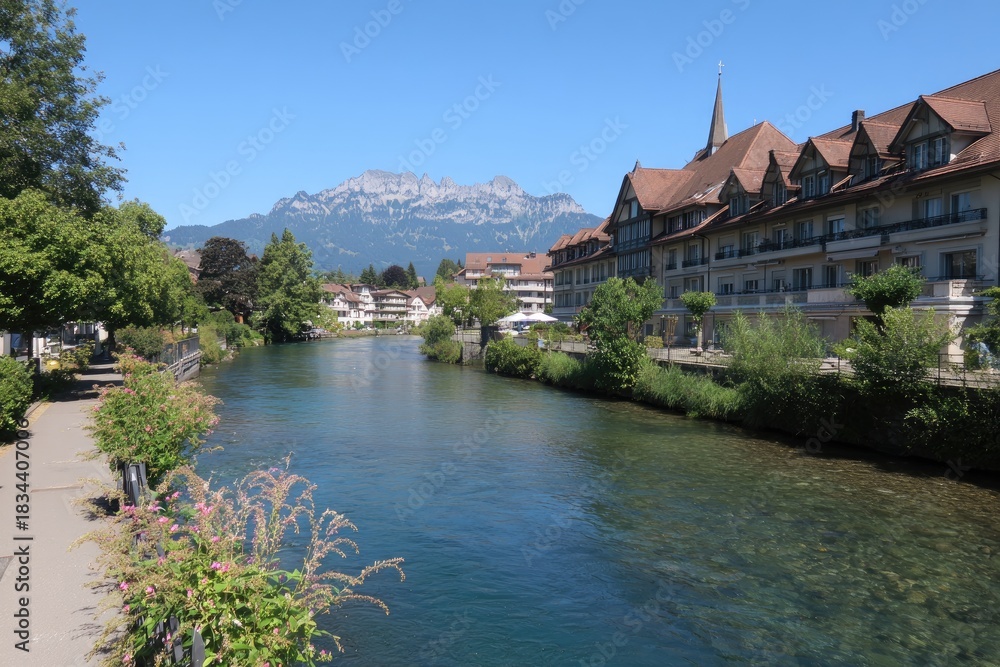 Fototapeta premium Riverfront view of buildings and mountains under a clear blue sky, summer